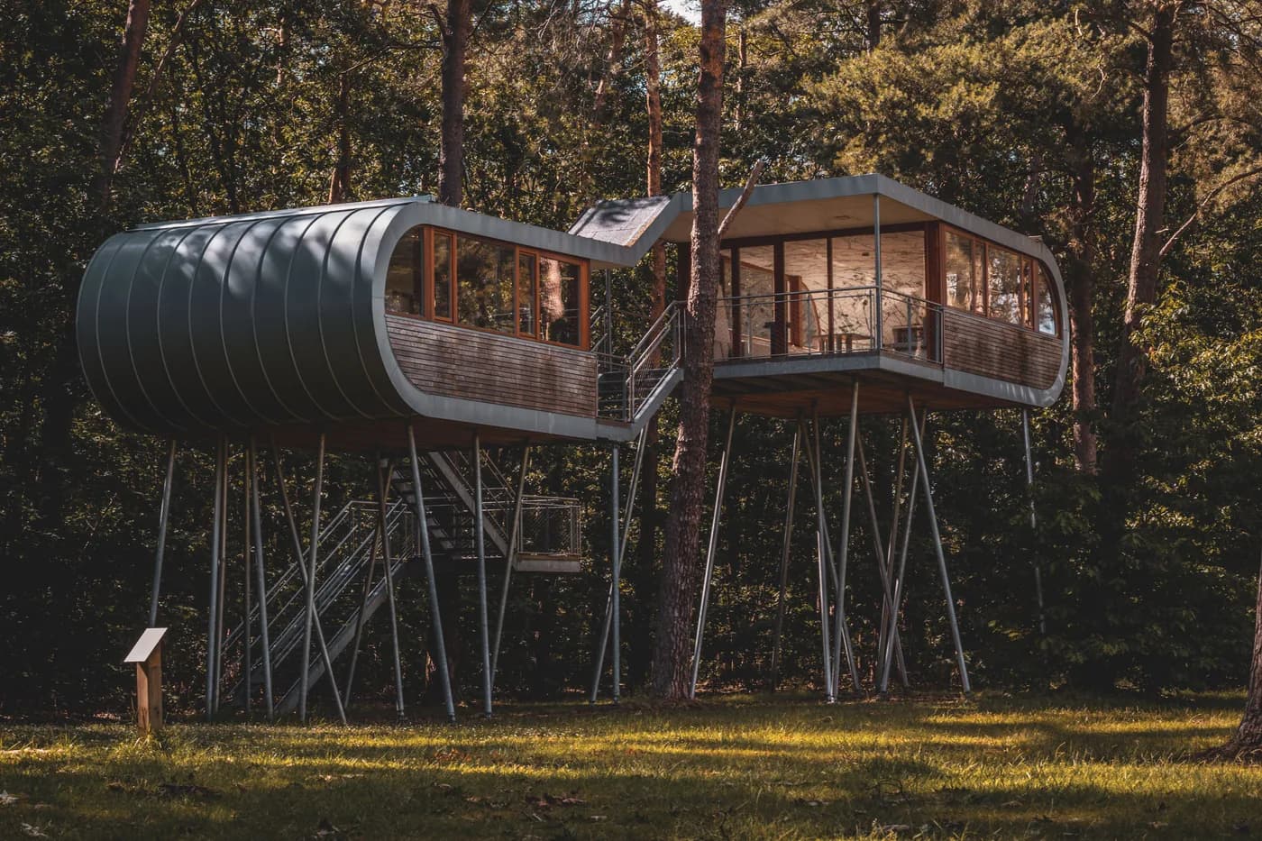 BookNoFees Imagem de exemplo: duas casas na árvore modernas, erguidas sobre pilares, com escadas de metal entre as árvores da floresta
