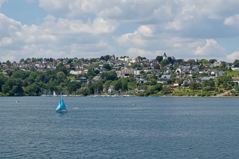 BookNoFees: Langscheid, às margens do Sorpesee, perto de Sundern: a vila à beira do lago, vista de cima, com um veleiro à frente