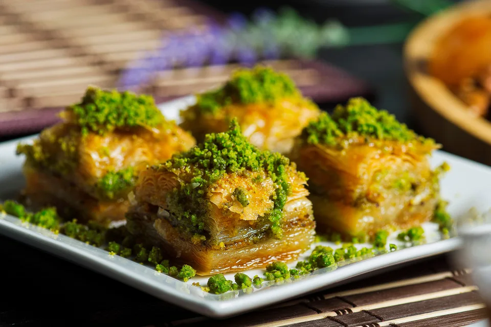 BookNoFees: Fotografia ilustrativa: Quatro pedaços de baklava em camadas com pistácios triturados num prato branco